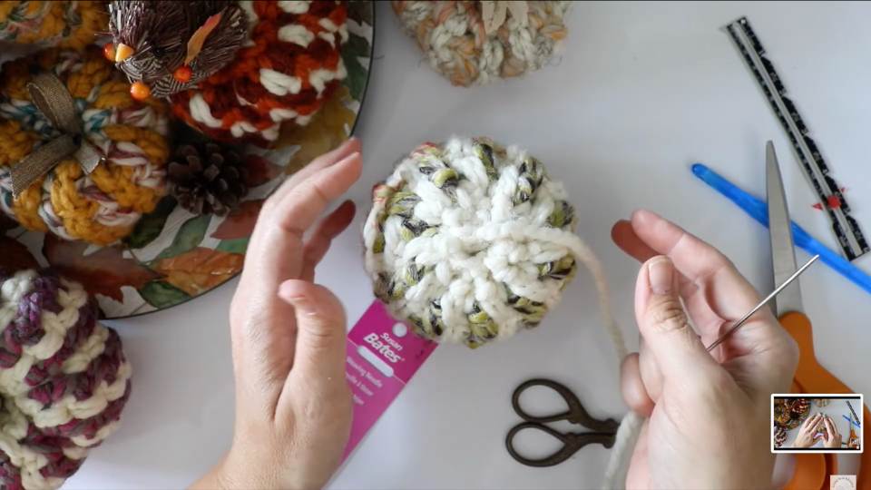 A person holds a chunky white and multicolored yarn pumpkin; scissors, needle, and more yarn pumpkins sit on the table nearby.