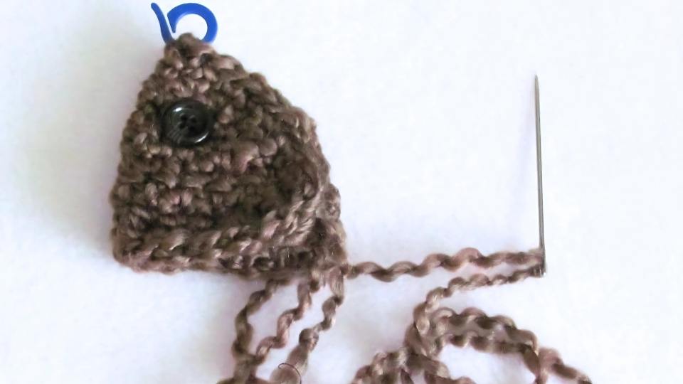 Brown crocheted fish with a black button eye, loose yarn ends, blue stitch marker, and sewing needle attached; no visible text. Free crochet pattern