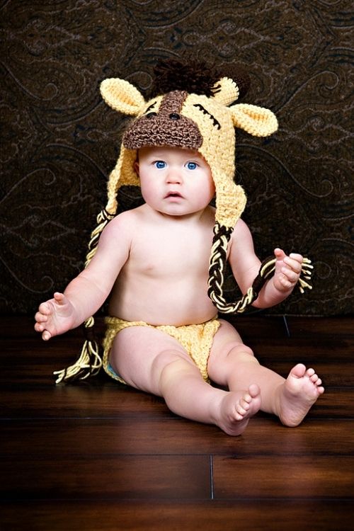 Baby with blue eyes wearing yellow crochet giraffe hat and diaper cover sits on wooden floor with dark patterned backdrop. Free crochet pattern