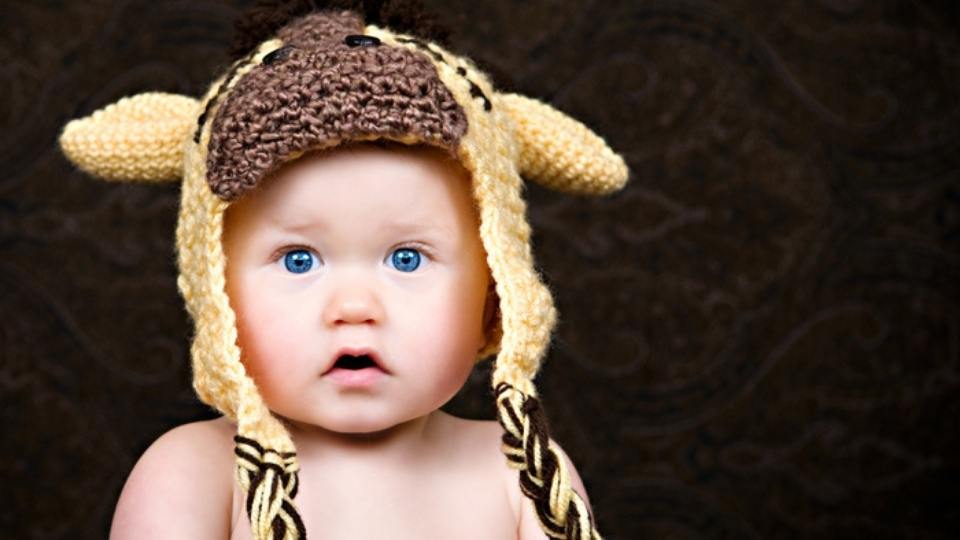 Baby with blue eyes in a yellow and brown giraffe crochet hat with ears and braided strings, set against a dark background. Free crochet pattern