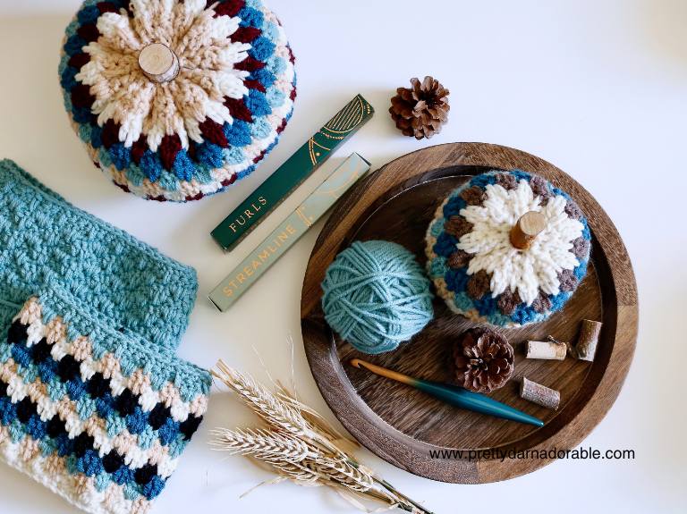 Top view of crocheted pumpkins, green yarn, crochet hooks, blue and cream crochet piece, pinecones, wheat, and corks on wood. Free crochet pumpkin pattern