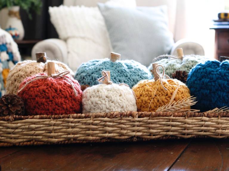 A woven tray on a wooden table holds soft, multicolored crochet pumpkins with short wooden stems; no visible text or promos. Free crochet pattern