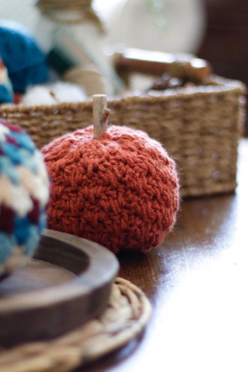 Small, round orange crochet pumpkin with a stick stem rests on a wooden table beside a woven basket and various handmade crafts. Free crochet pattern