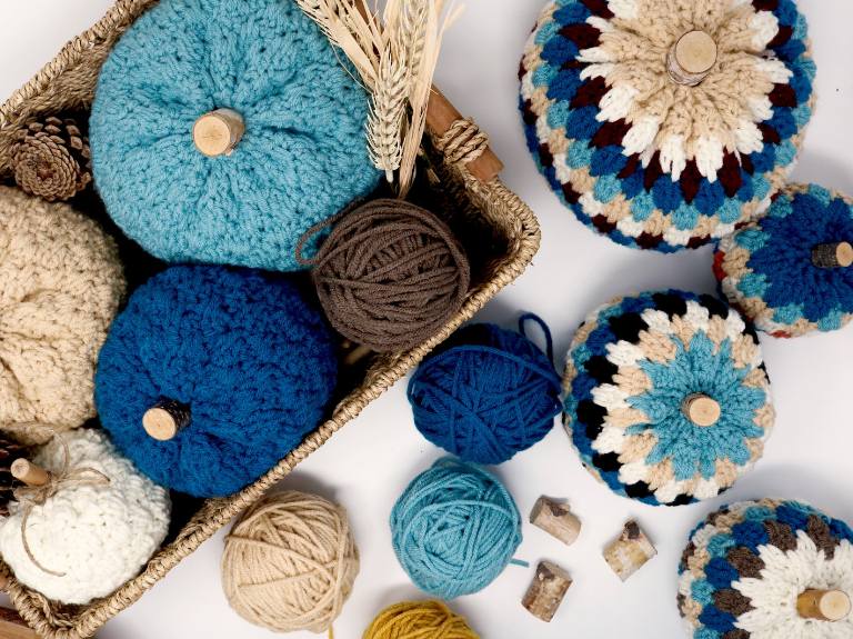 Woven basket with blue, cream, and teal crochet pumpkins, surrounded by yarn balls, pinecones, and corks on a white surface. Free crochet pattern