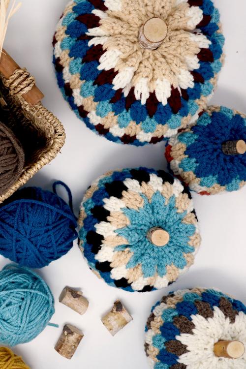 Crocheted pumpkins in bright colors with wooden stems sit on a white surface with yarn balls, crochet hook, tray, pinecones, and corks. Free crochet pattern