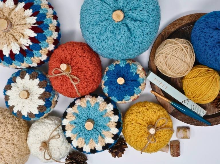 Crocheted pumpkins in bright colors with wooden stems sit on a white surface with yarn balls, crochet hook, tray, pinecones, and corks. Free crochet pattern