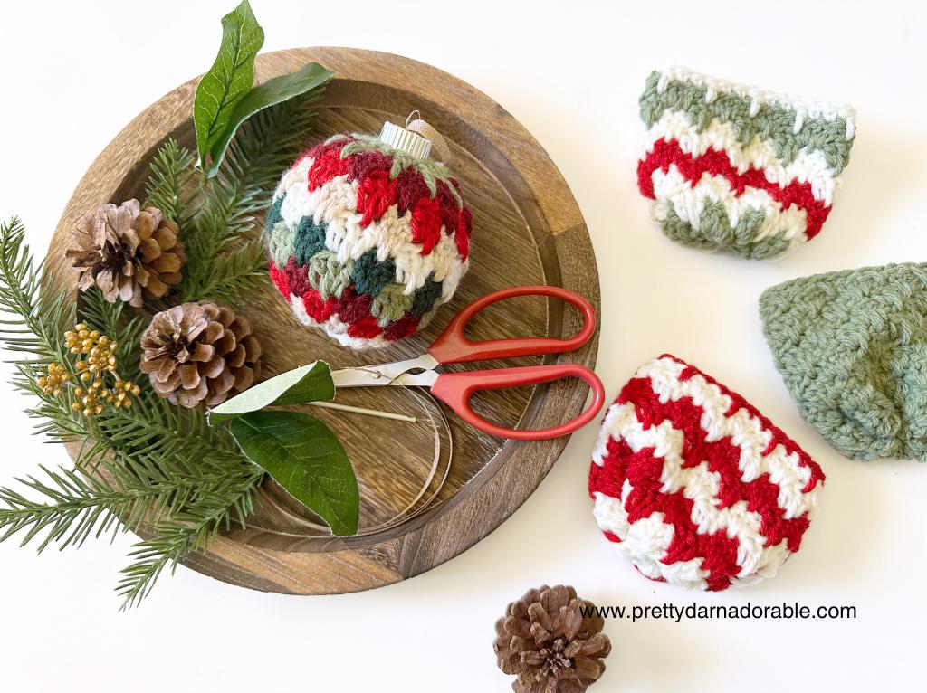 Wooden tray with red scissors, a crochet ornament, pine cones, and greenery; red, white, and green crochet pouches nearby. Free easy crochet Christmas Bauble pattern