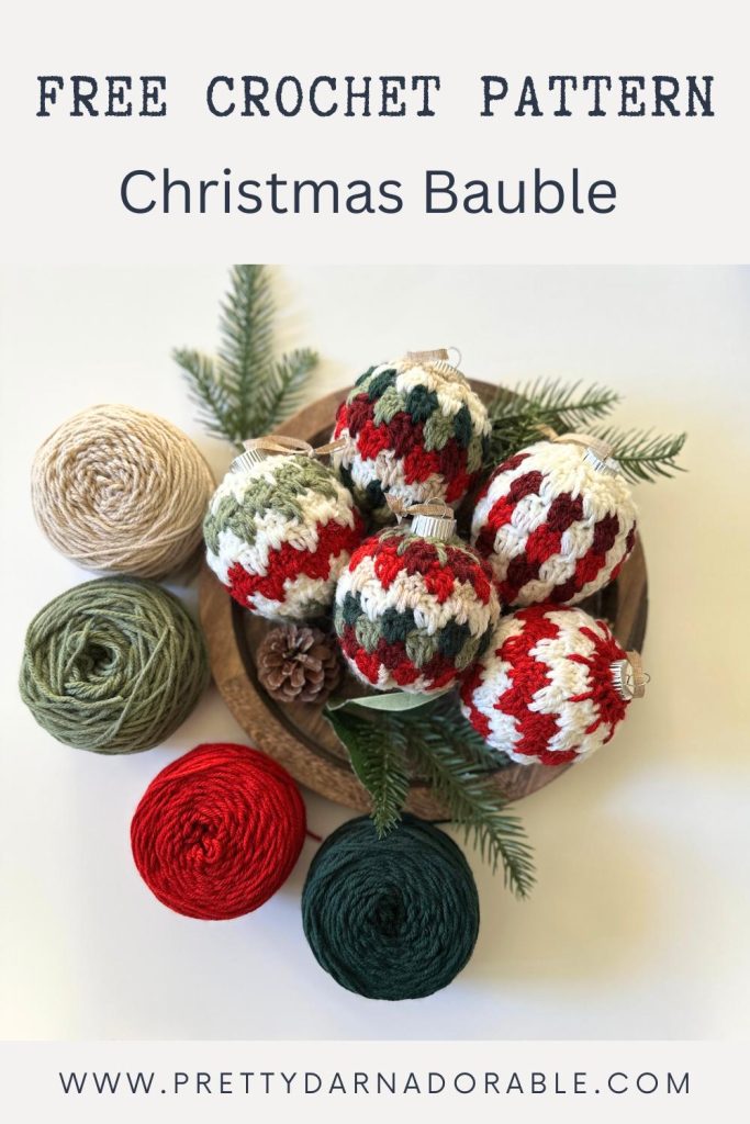 A wooden bowl holds red, green, and cream crocheted Christmas baubles, with yarn balls and pine branches nearby. Text: Free Crochet Pattern Christmas Bauble. Free crochet pattern