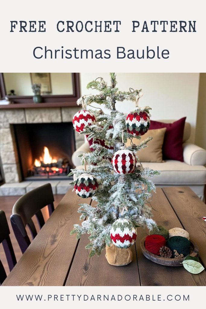Small snowy Christmas tree with crochet baubles on a wood table, yarn and tools nearby, cozy décor, fireplace and sofa behind. Free easy crochet Christmas bauble pattern