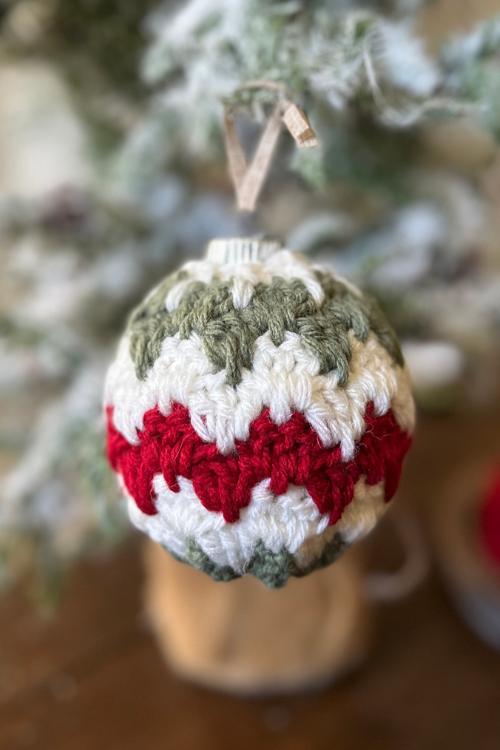 A green, white, and red striped crocheted ornament hangs from a snow-dusted Christmas tree branch. Free easy crochet Christmas Bauble pattern