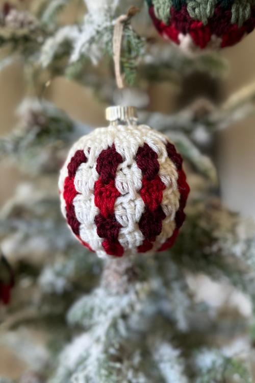 Close-up of a Christmas ornament with a red, white, and dark green crocheted cover on a snow-dusted tree branch. No visible text. Free easy crochet Christmas Bauble pattern