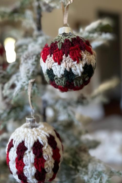 Two red, white, and green crocheted Christmas ornaments hang on a snow-dusted tree with a softly blurred background. Free easy crochet Christmas Bauble pattern