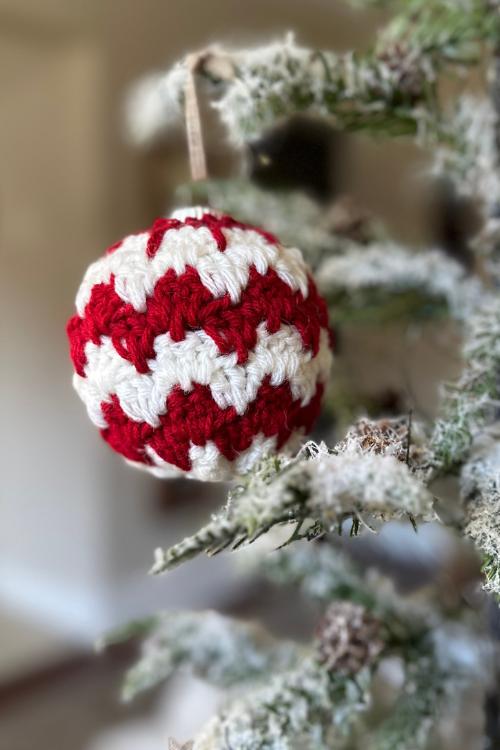 Close-up of a snowy Christmas tree branch with a red and white crochet ornament on a ribbon; background is blurred. Free easy crochet Christmas Bauble pattern