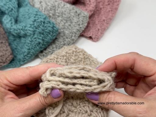 Hands with purple nail polish hold a folded beige fabric; blue, pink, and beige textured items appear in the background. Free crochet pattern