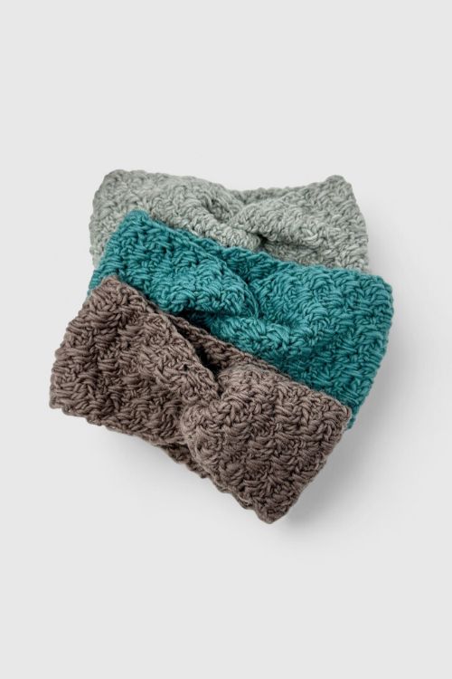 Three crocheted headbands in light gray, teal, and brown are stacked on a plain background, each with a center twisted knot. Free crochet pattern