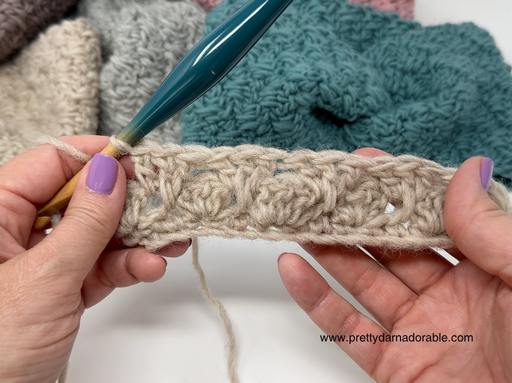 Two hands with purple nail polish hold a beige crochet project and teal hook, with colorful finished pieces in the background. Free crochet pattern