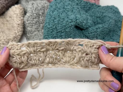 Hands with lavender nails hold a beige crochet swatch with bobble pattern; gray and teal crochet pieces are in the background. Free crochet pattern