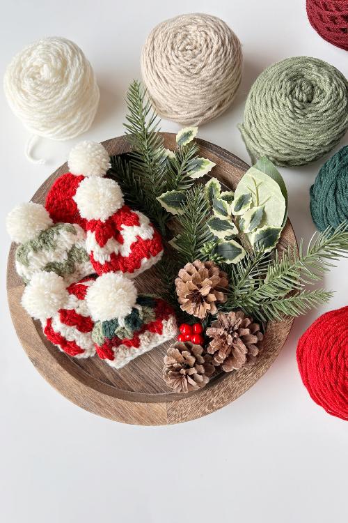 A wooden plate with three red and white mini hats with pom-poms, pinecones, berries, foliage, and yarn balls in soft colors. Free crochet mini hat christmas ornament pattern