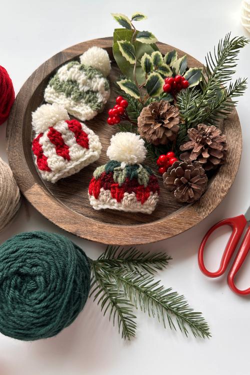 A wooden plate holds three small wool hats, pinecones, holly, and red berries amid yarn balls, pine branches, and red scissors.Free crochet mini hat christmas ornament pattern