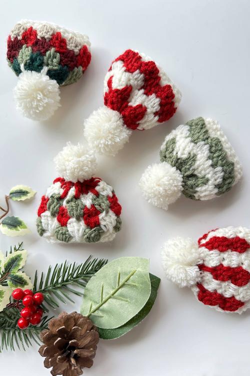 Five small winter hats with white pom-poms in red, green, and white patterns sit on a white surface beside holiday greenery. Free crochet mini hat christmas ornament pattern