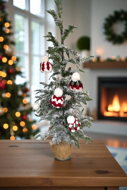 Small snowy potted Christmas tree with red and white hat ornaments on a wooden table; fireplace, wreath, and lit tree in background. Free crochet mini hat christmas ornament pattern