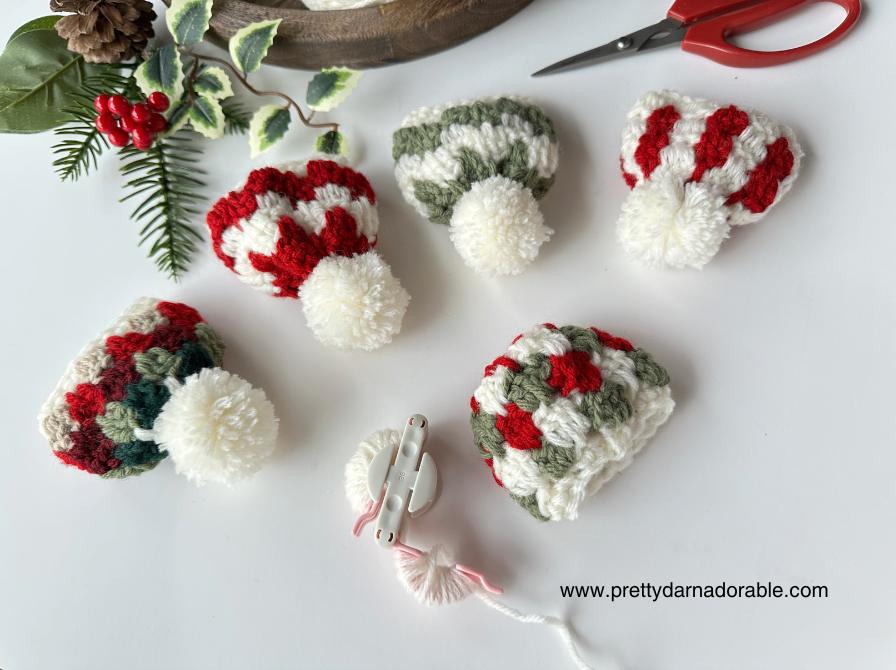 Five small crocheted hats in red, white, and green on a white surface with greenery, scissors, pom-pom maker; www.prettydarnadorable.com at bottom. Free crochet pattern