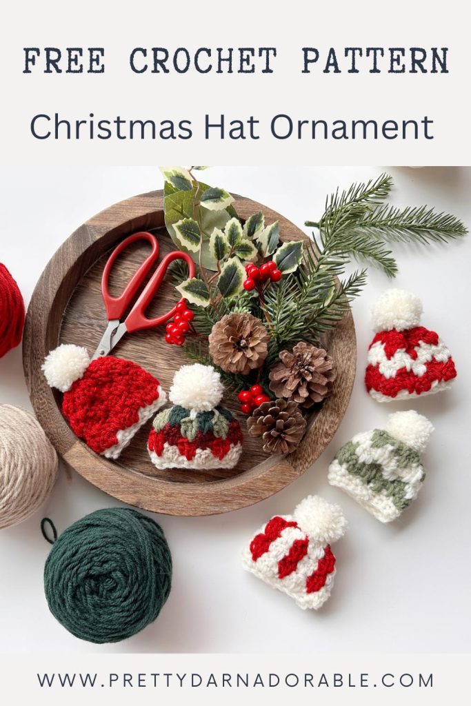 Round wooden tray with pine cones, greenery, red scissors, and four crocheted Christmas hats; yarn and “Free Crochet Pattern” text visible. Free crochet mini hat christmas ornament pattern