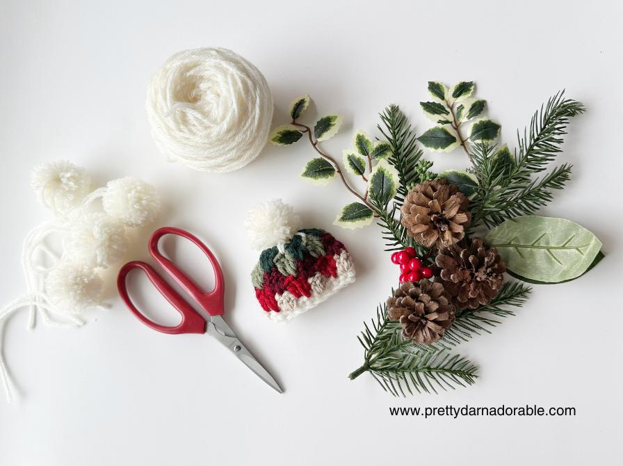 Craft supplies on a white surface: white yarn, pom-poms, red scissors, a small crocheted hat, pinecones, berries, and greenery. Free crochet mini hat christmas ornament pattern