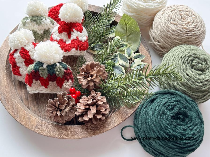 Wooden tray with small crocheted red, white, and green hats, pinecones, greenery, berries; 3 yarn balls sit nearby.Free crochet mini hat christmas ornament pattern