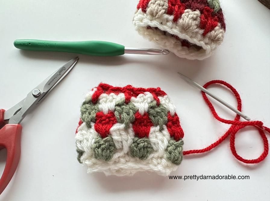 Two red, green, and white crochet granny squares with a green hook, red scissors, metal needle on white; Free crochet mini hat christmas ornament pattern