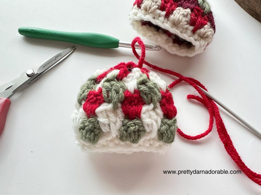 A red, green, and white crocheted ornament in progress rests on a white surface with tools and www.prettydarnadorable.com visible. Free crochet mini hat christmas ornament pattern