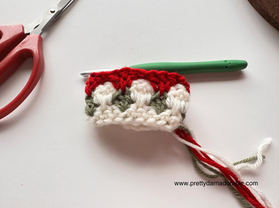 Red, white, and green yarn crochet piece with a green hook and red scissors on white surface;