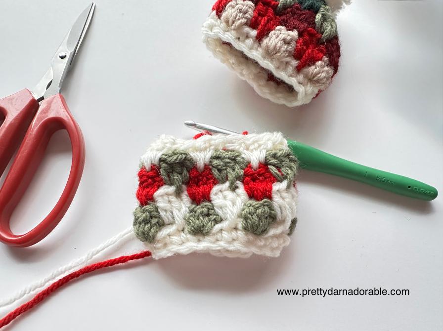 Green, red, and white yarn with a crochet hook, red scissors, and a finished piece on a white surface; www.prettydarnadorable.com shown.