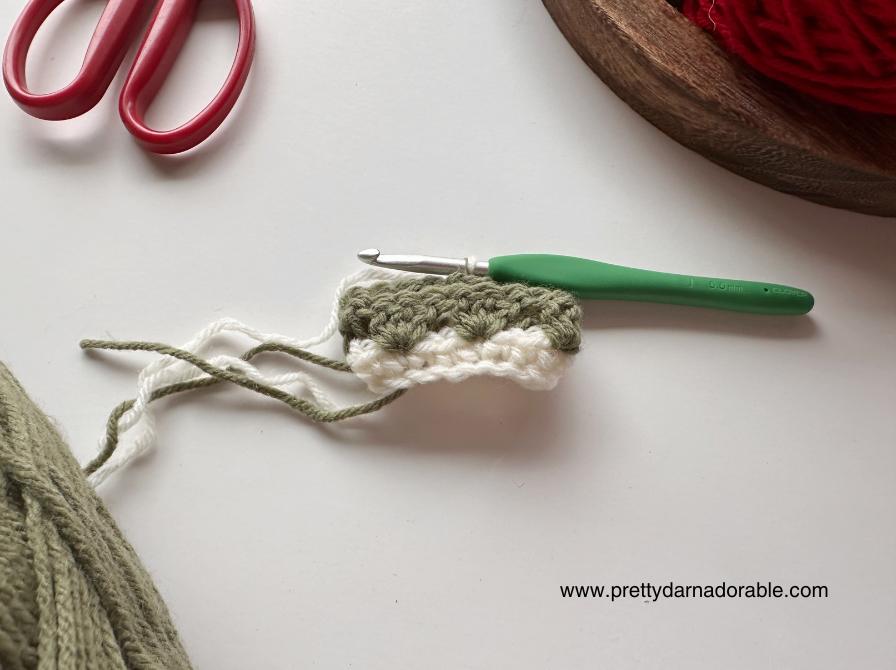 A green-handled crochet hook in a green and white swatch, red scissors, yarn ball, and www.prettydarnadorable.com on white surface. Free crochet mini hat christmas ornament pattern