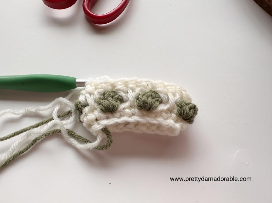 White and green crochet project with bobble stitches, green crochet hook, red scissors, and a visible website URL in the background. Free crochet pattern