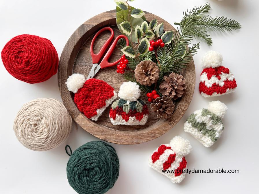 A wooden tray with small yarn hats, pinecones, scissors, faux pine, and red berries is surrounded by red, cream, and green yarn balls. Free crochet mini hat christmas ornament pattern