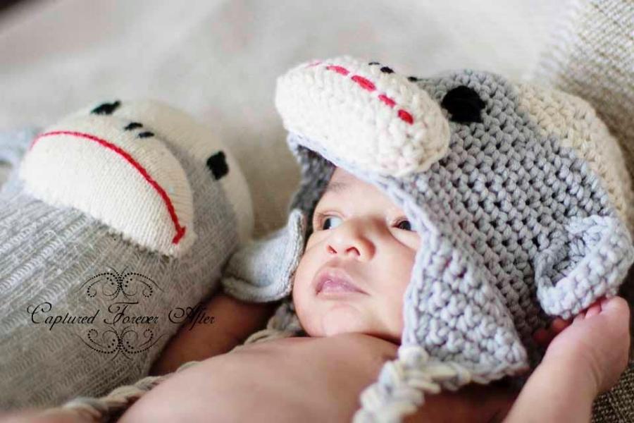A newborn in a gray crochet monkey-face hat is cradled in hands beside a matching gray sock monkey plush toy. No text visible. Free crochet pattern