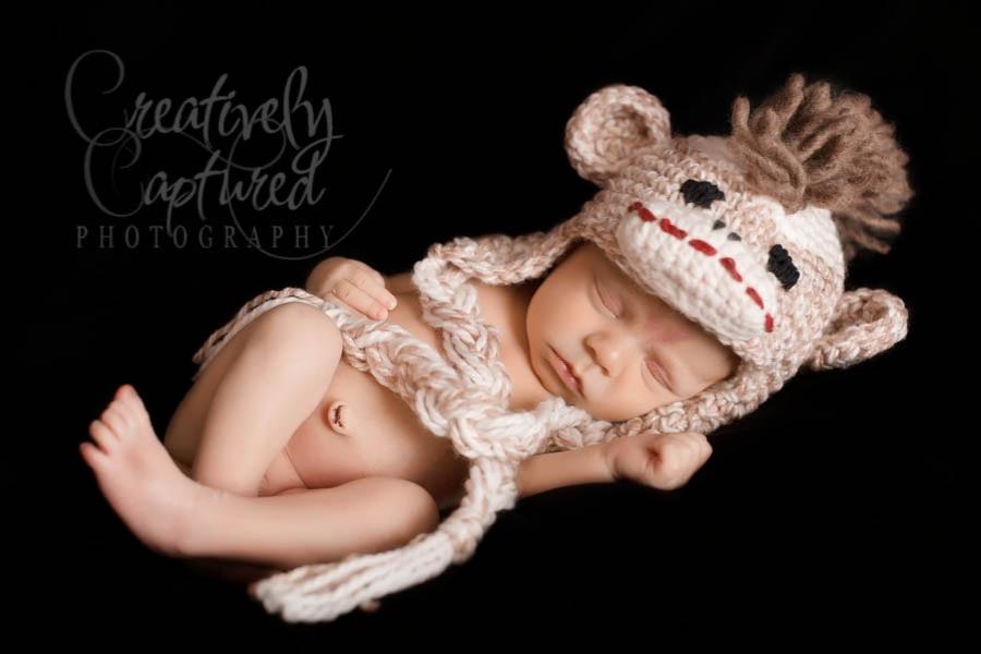 A newborn baby sleeps on a black background, wearing a sock monkey-themed hat with ear flaps and matching yarn swaddle. Free crochet pattern