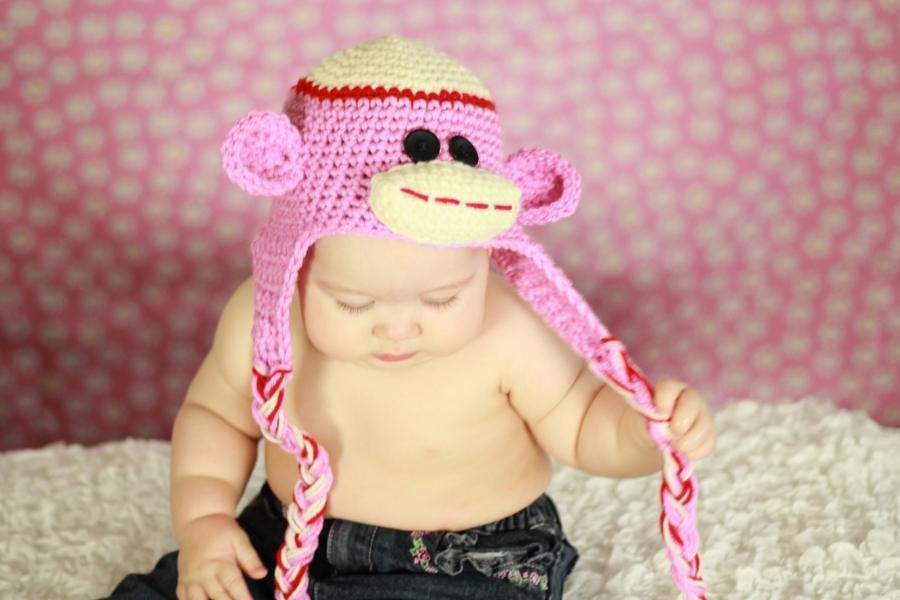 Baby in a pink crocheted monkey hat with ear flaps sits on a white blanket, pink polka dot background, holding a braided tassel. Free crochet pattern