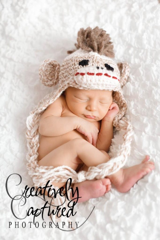 Newborn baby sleeps on a white textured blanket, wearing a sock monkey-style hat. "Creatively Captured Photography" logo bottom left. Free crochet pattern