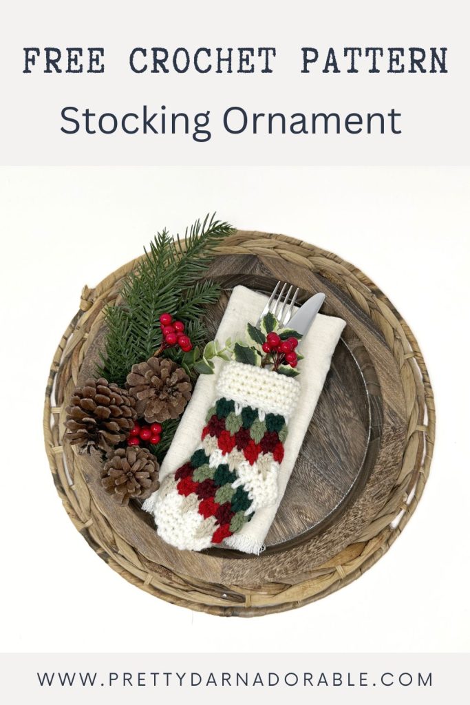 Small red, green, and white crocheted Christmas stocking holds silverware on napkin, on wooden plate with pinecones and berries. Text: Free Crochet Pattern Stocking Ornament. Free crochet pattern
