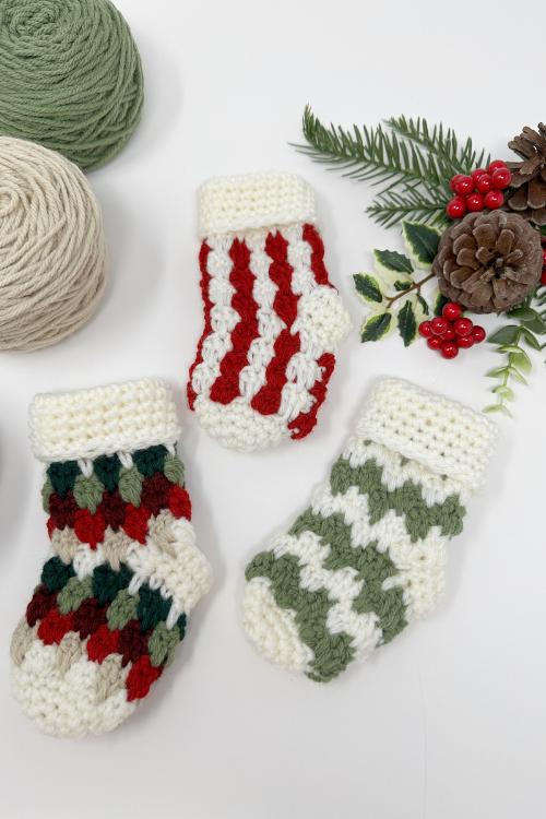Three small crocheted Christmas stockings in red, green, and white sit on a white surface with yarn balls and pine, cones, and berries. Free crochet pattern