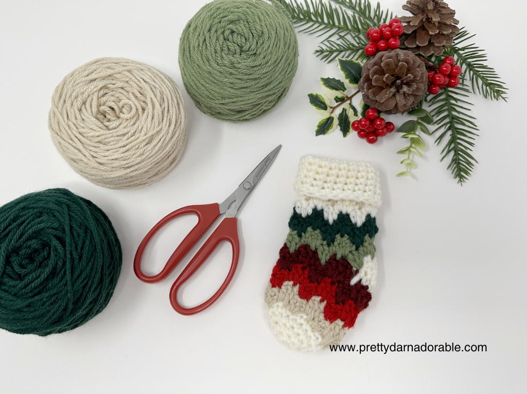 Cream, green, and dark green yarn balls, red scissors, a crocheted Christmas stocking, and pine decor on white. www.prettydarnadorable.com. Free crochet pattern