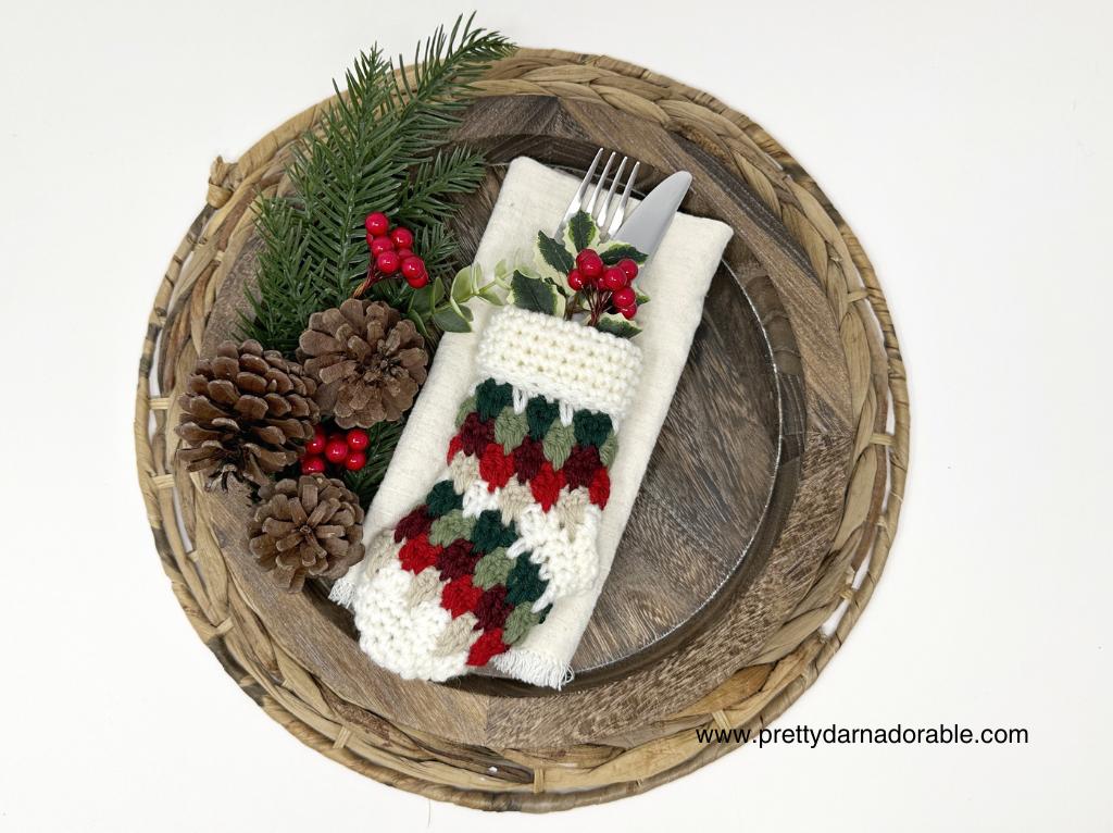 Festive holiday table setting with pinecones, greenery, red berries, and utensils in a crocheted red, green, and white holder; website text in corner. Free crochet pattern
