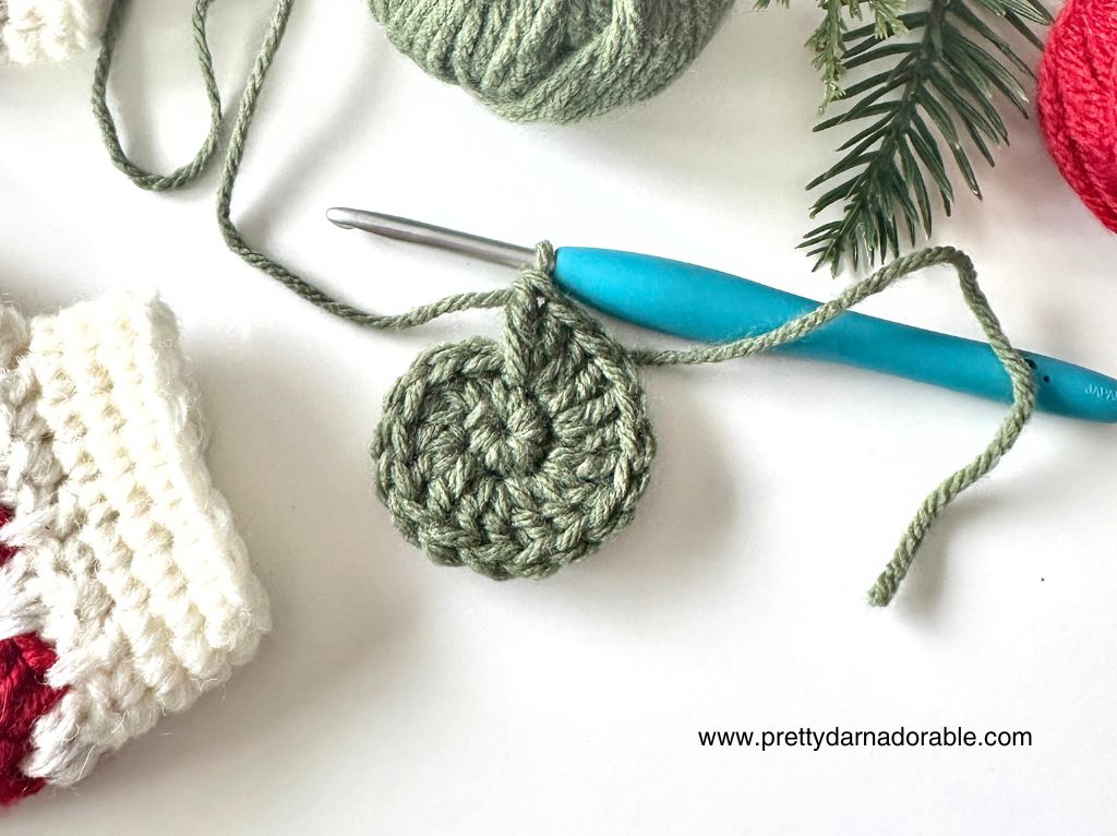 A blue crochet hook and green yarn make a half-finished circle with yarn balls, a stocking, pine branch, and www.prettydarnadorable.com. Free crochet pattern