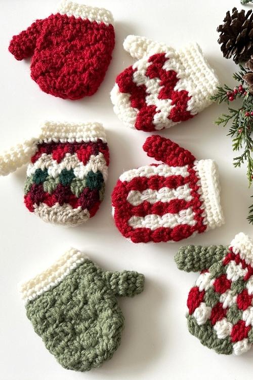 Six red, white, and green crocheted mittens lie on a white surface beside pinecones and evergreen branches for a festive look. Free crochet pattern