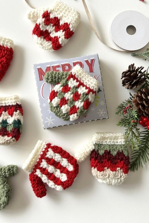 Crocheted mini mittens in red, white, and green with a "Merry Christmas" sign, ribbon spool, pinecones, and greenery on white. Free crochet pattern