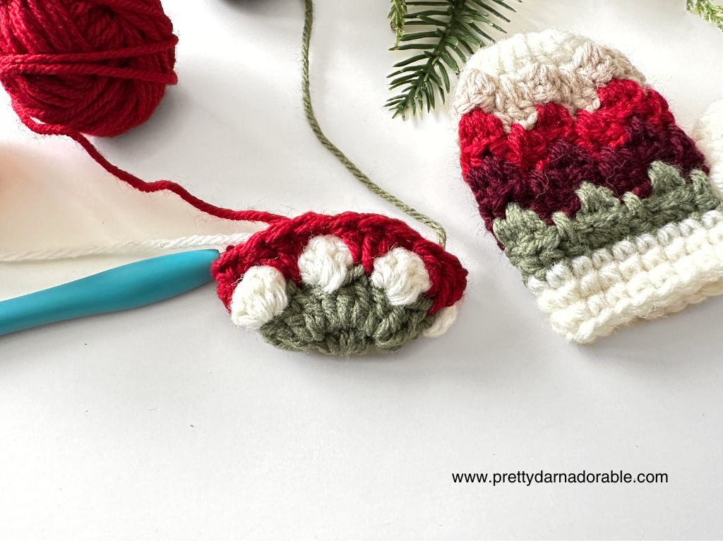 Red, white, and green crochet mitten beside a project in progress, blue hook, red yarn ball, evergreen branches; www.prettydarnadorable.com. Free crochet pattern