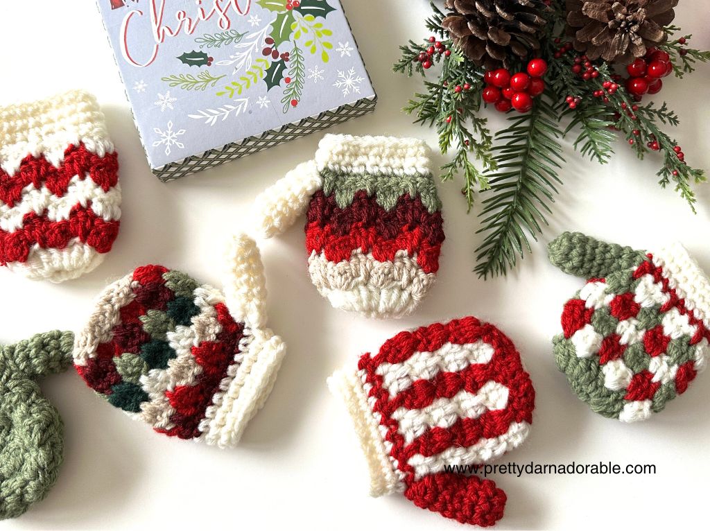 Red, green, and white crocheted Christmas mittens with varied patterns rest on a white surface beside a notepad, greenery, and pinecones. Free crochet pattern