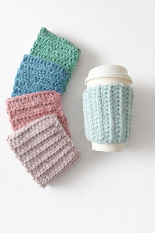 Four textured cup sleeves in green, blue, pink, and gray beside a white takeaway cup with a light blue sleeve on a white background. Free crochet pattern